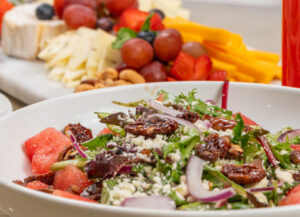 A bowl of mixed green salad with red onions, crumbled cheese, and tomatoes, with a platter of sliced cheese, fruits, and nuts in the background.