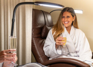 A woman in a white robe sits in a reclining chair, smiling and holding a glass of juice under a reading light. Another person holds a glass of sparkling water nearby.