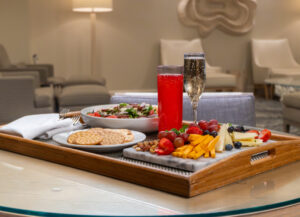 A wooden tray on a table holds a salad, crackers, a cheese and fruit platter, a red drink, and a glass of sparkling wine in a modern lounge setting.