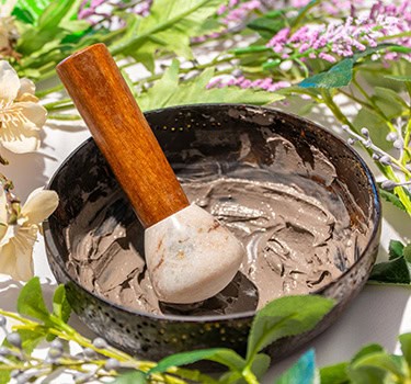 A wooden-handled pestle rests in a bowl containing a grayish clay mixture, surrounded by green leaves and light purple and yellow flowers.