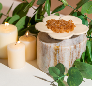 A flower-shaped dish with brown sugar scrub sits on a marble container next to three lit candles and green leafy branches.