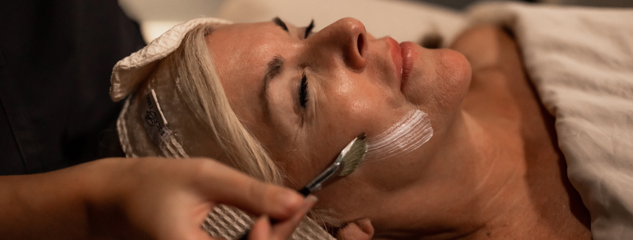 A woman with blonde hair lies down with eyes closed while a brush is used to apply a white facial cream to her cheek in a spa setting.