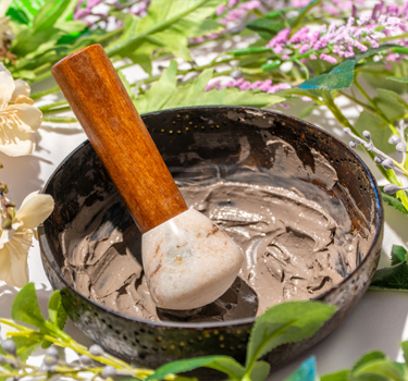 A bowl filled with gray clay, featuring a small wooden-handled mixing tool, is surrounded by green leaves and purple and white flowers.